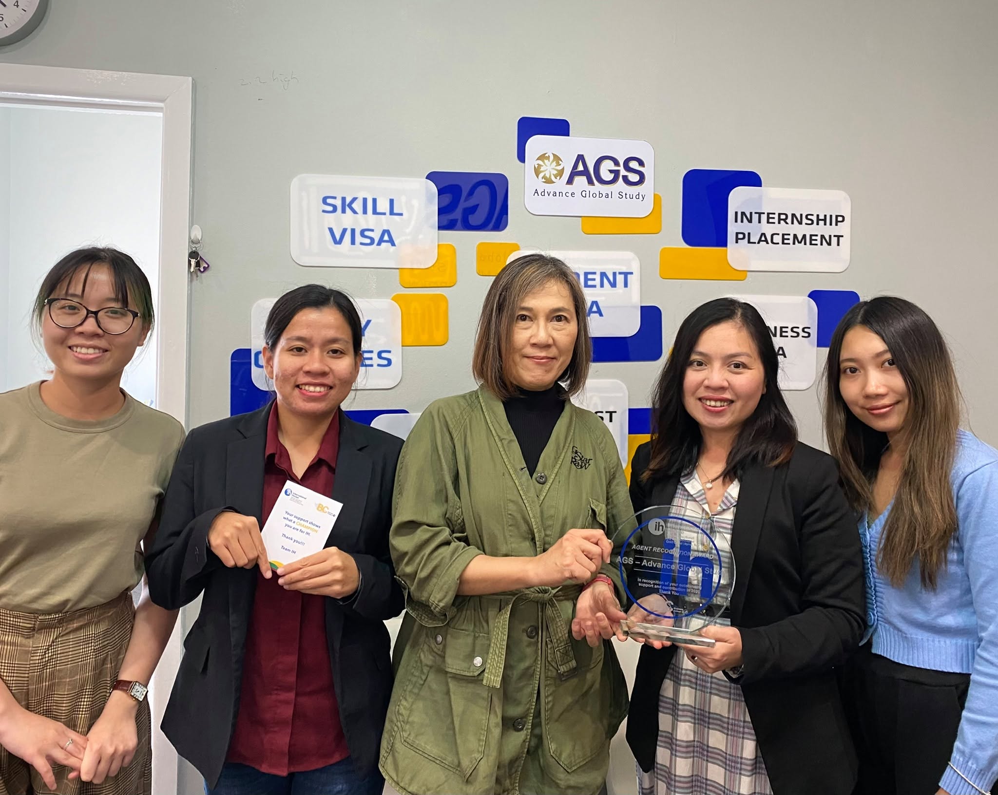 Five team members smiling in front of the AGS wall display, holding an agent recognition award